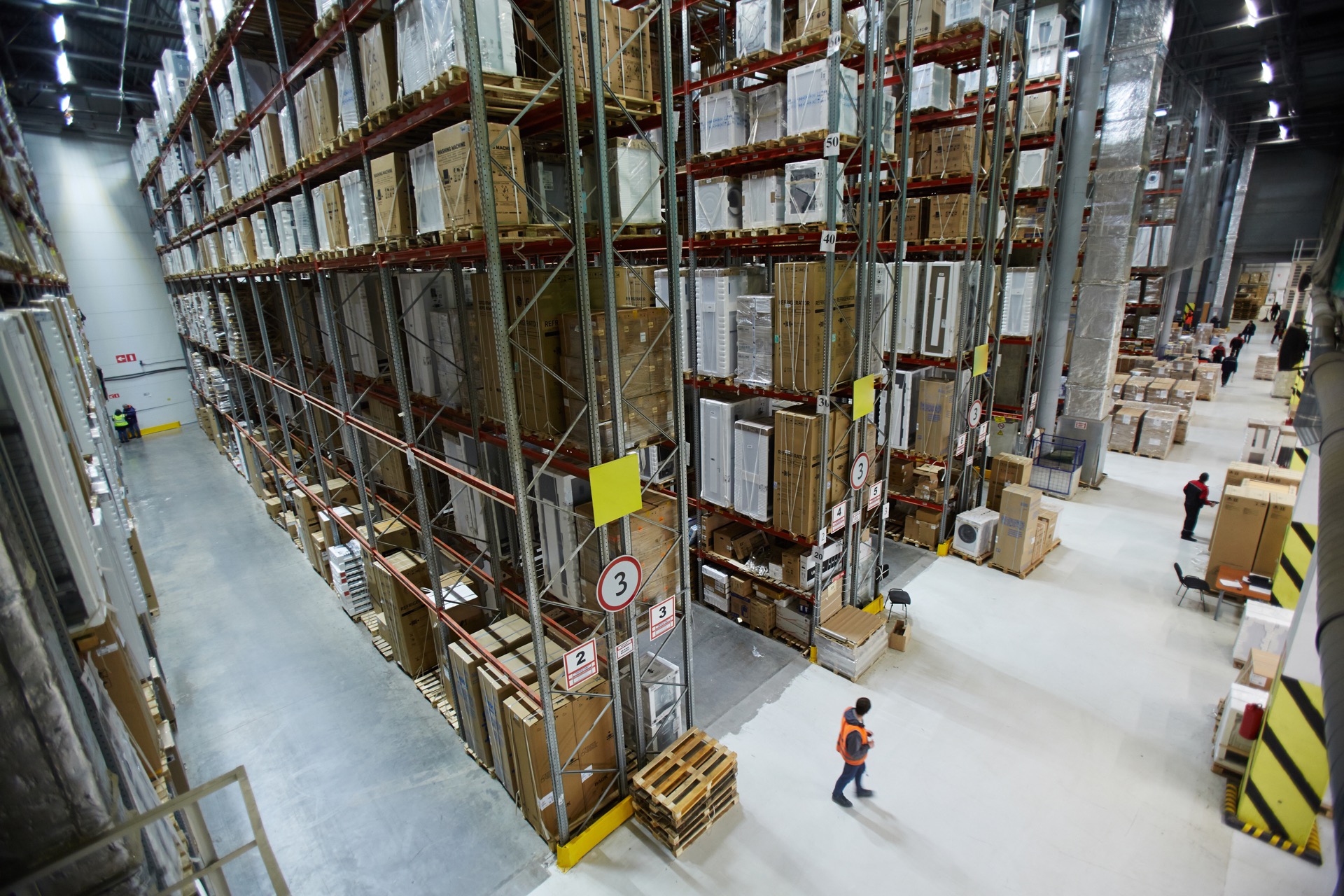 Large warehouse with tall storage racks and a worker in an orange vest walking the main aisle.